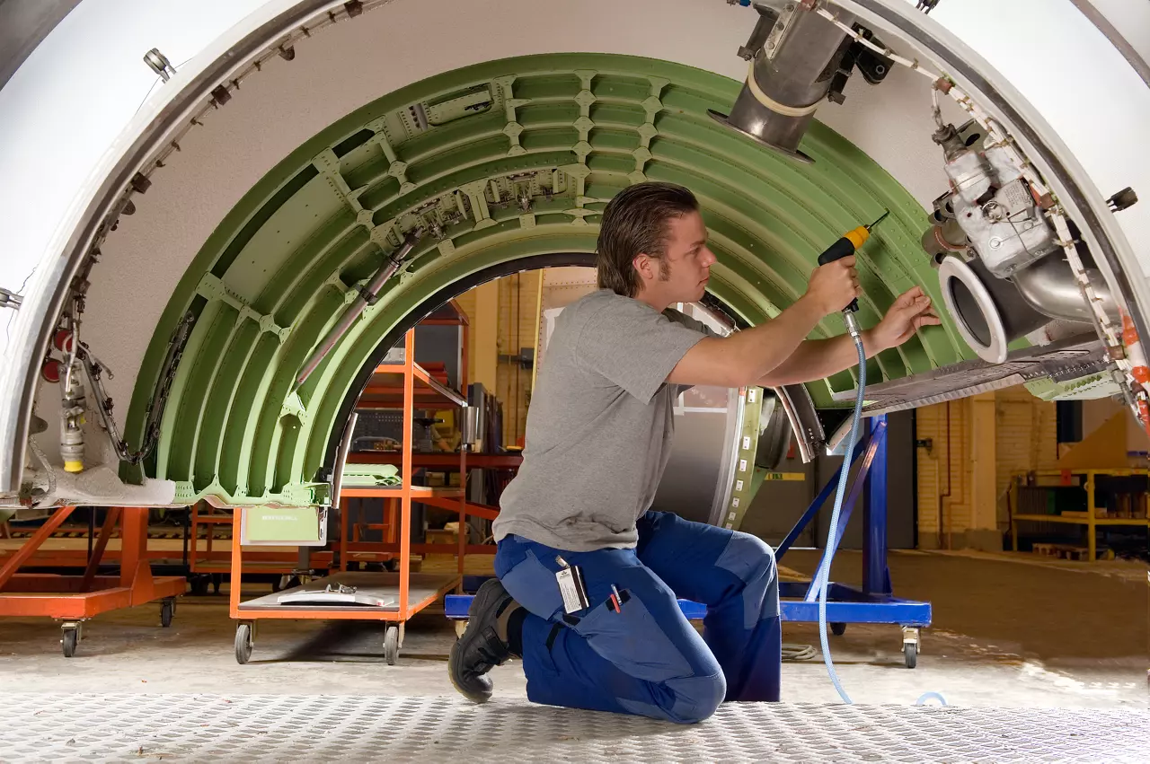 Technician using an air-powered tool on an aircraft fuselage section in an assembly hall